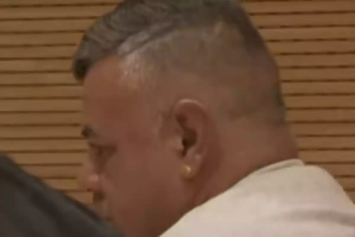 Un hombre de perfil con cabello corto y canoso, usando un pendiente en la oreja izquierda, en un entorno con fondo de madera.