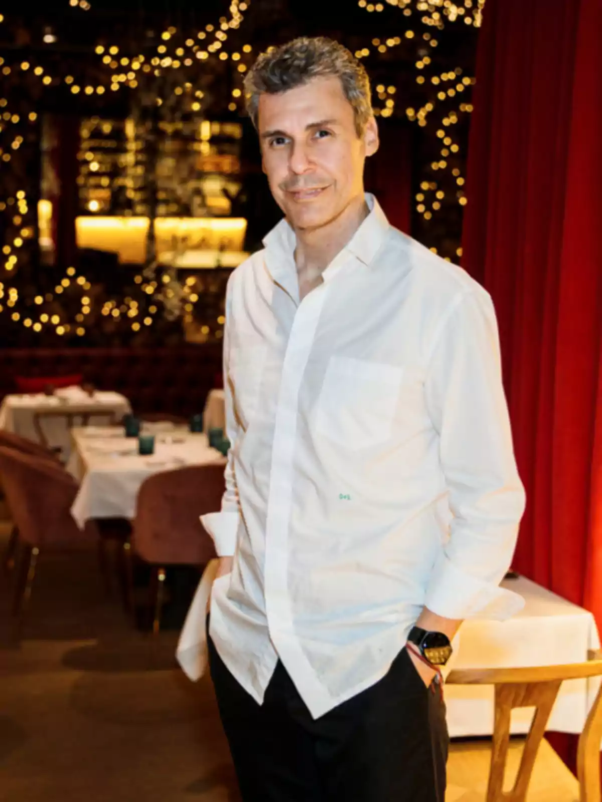 Un hombre con camisa blanca y reloj negro posa en un restaurante decorado con luces.
