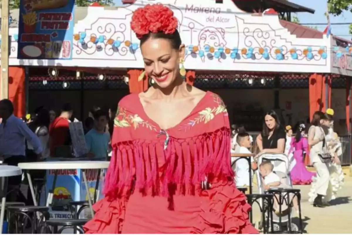 Mujer vestida con traje tradicional flamenco rojo y flores en el cabello, sonriendo en una feria con personas y puestos de comida al fondo.