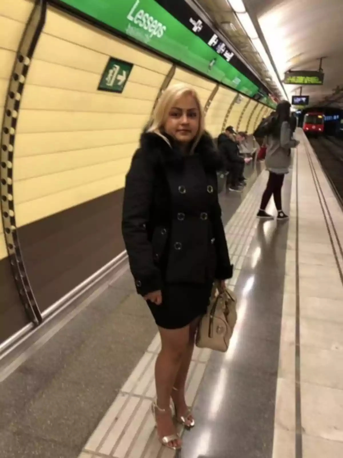 Una mujer con cabello rubio y abrigo negro está de pie en una estación de metro llamada Lesseps, sosteniendo un bolso beige.