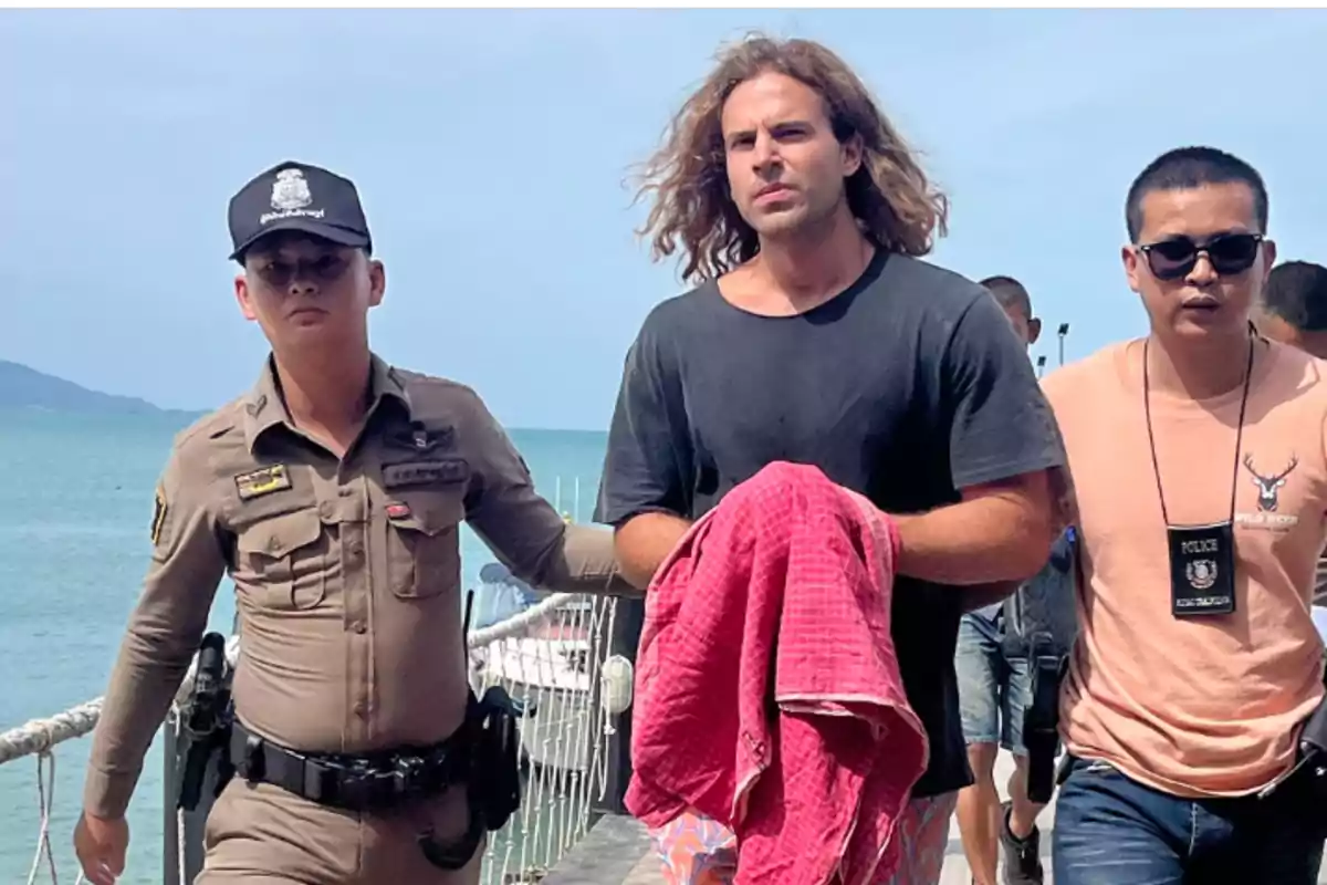 Un hombre escoltado por dos oficiales de policía en un muelle con el mar de fondo.