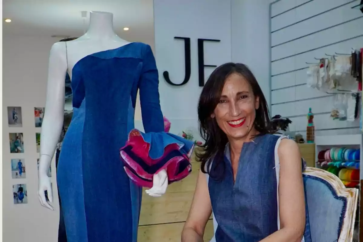 Mujer sonriente sentada junto a un maniquí con un vestido de mezclilla en una tienda de moda.