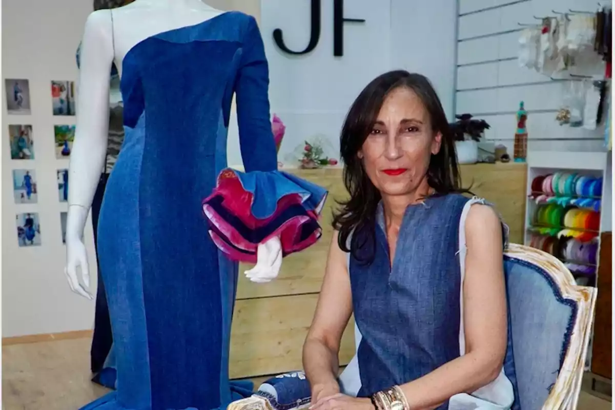 Una diseñadora de moda posa junto a un maniquí que lleva un vestido de mezclilla azul con un diseño asimétrico y detalles en las mangas.