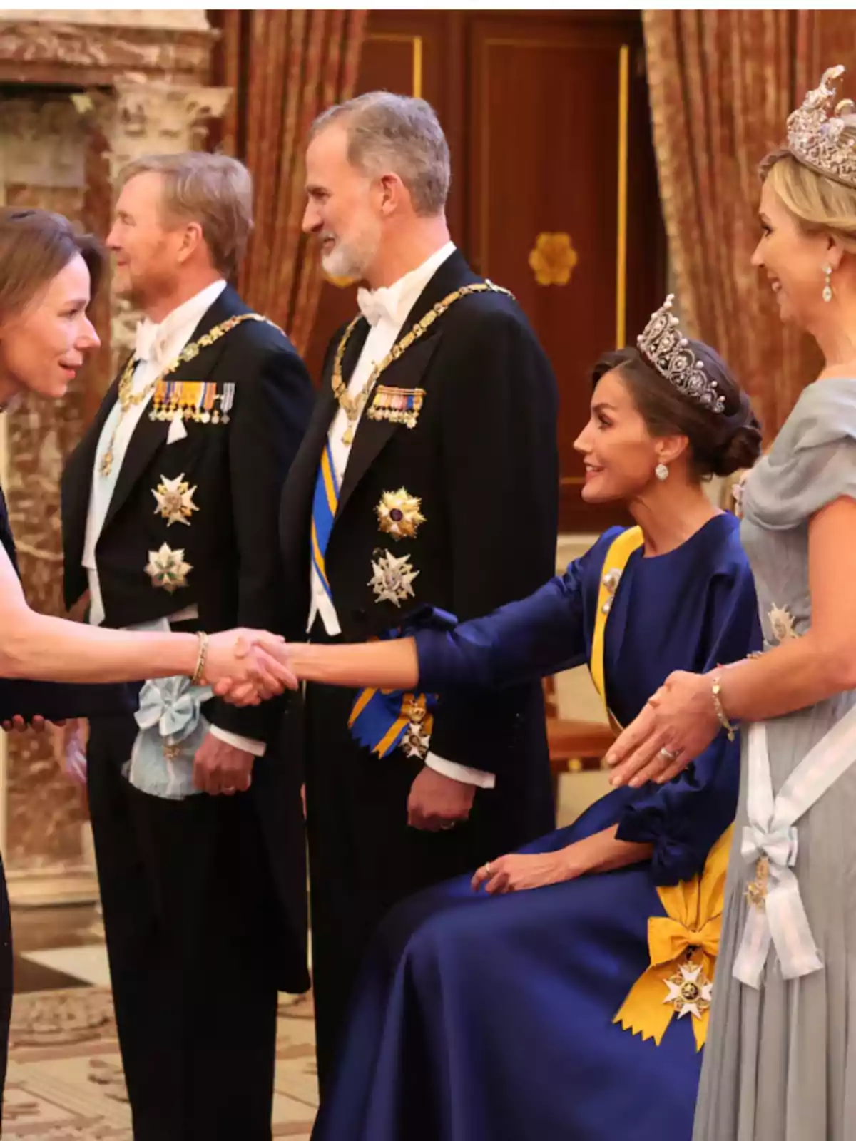 Personas vestidas de gala, algunas con tiaras y medallas, en un evento formal, una de ellas estrechando la mano de otra.