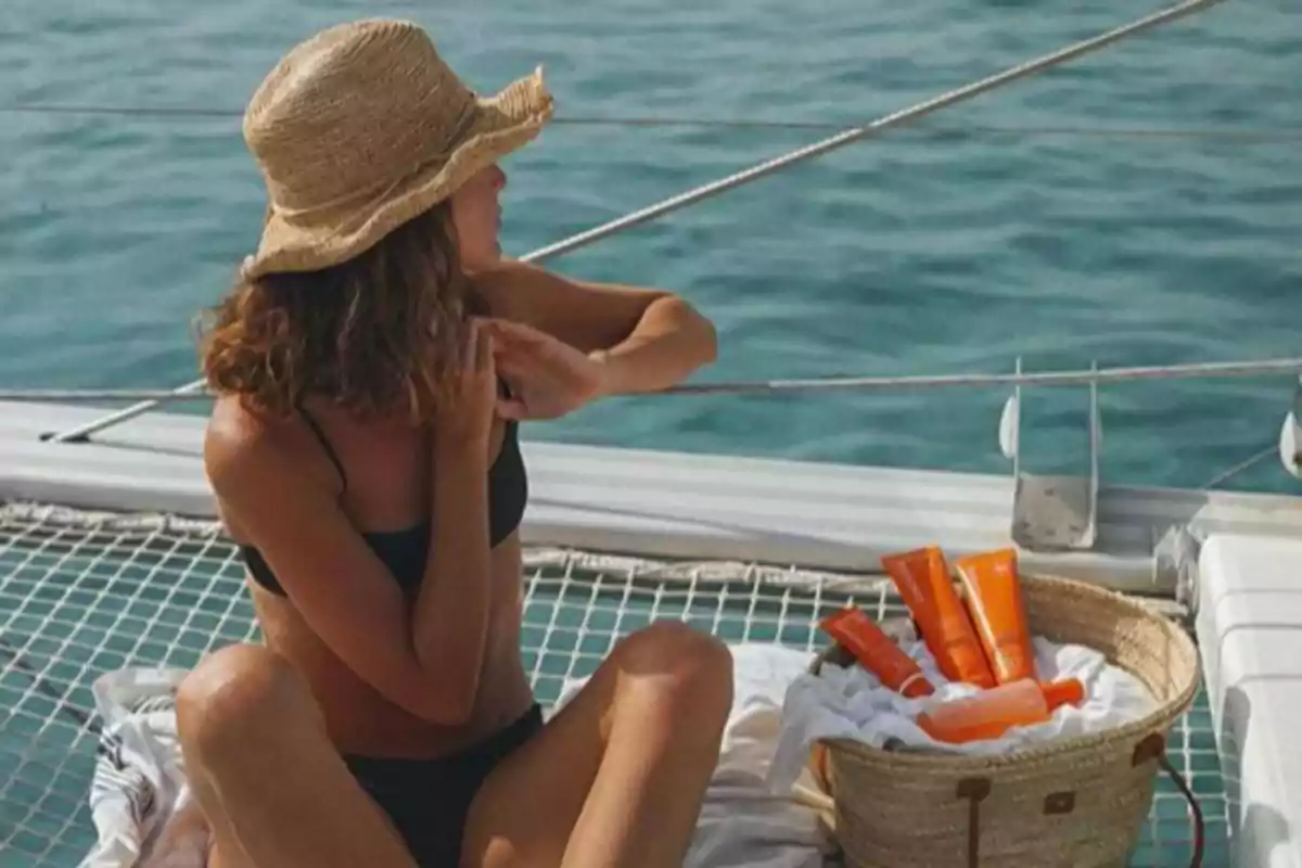 Persona en traje de baño y sombrero de paja sentada en la cubierta de un barco aplicándose protector solar con un cesto lleno de productos solares a su lado y el mar de fondo.