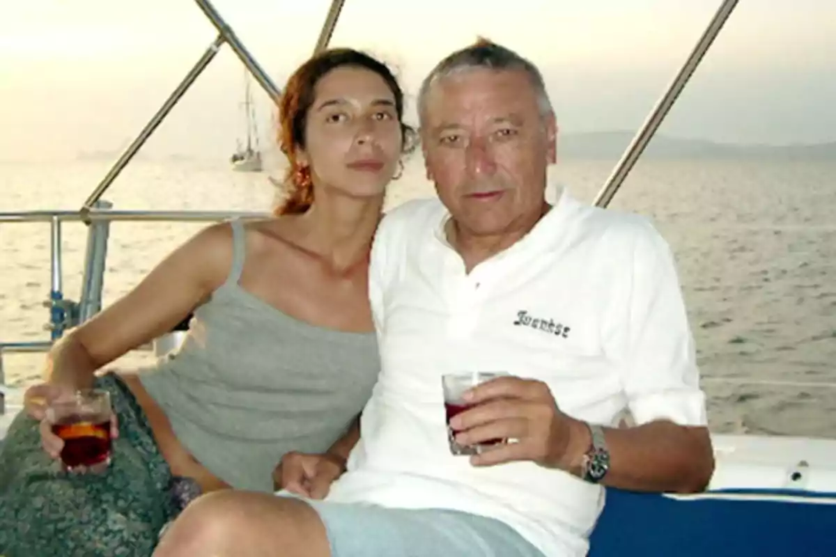 Una pareja sentada en un barco disfrutando de bebidas con el mar y un velero de fondo.