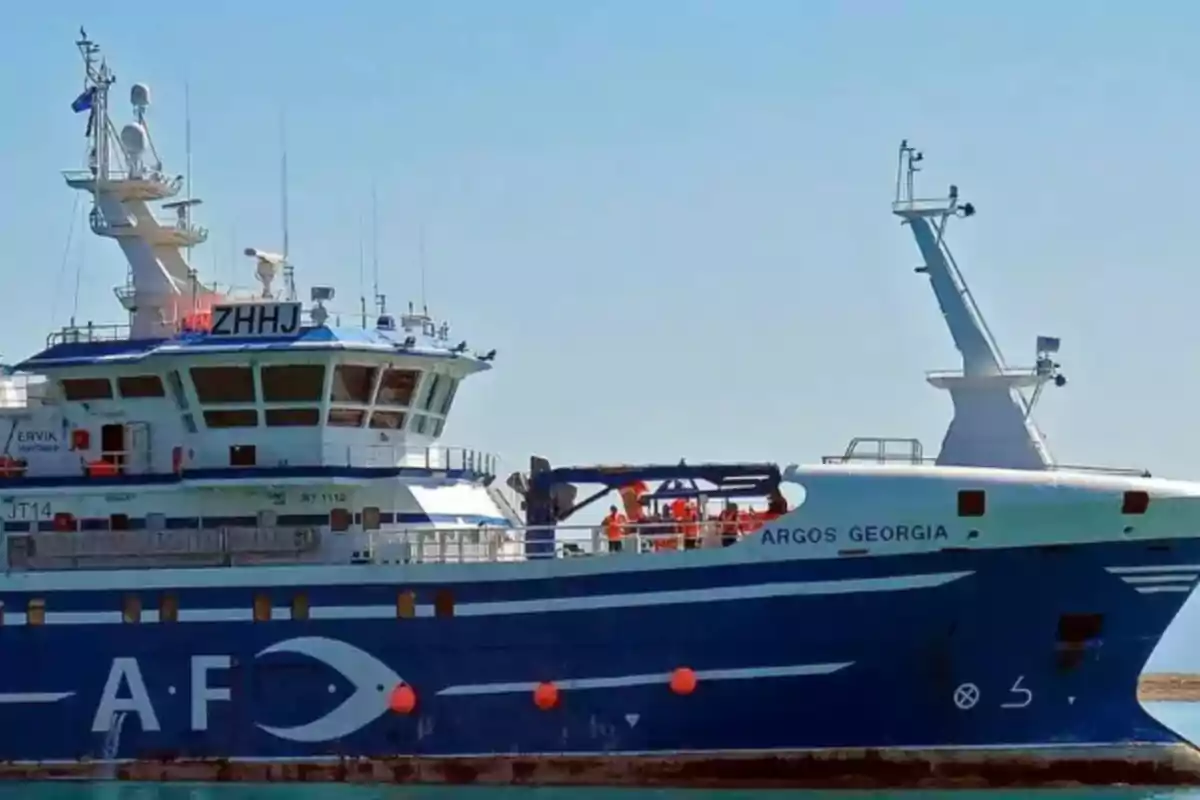 Un barco azul y blanco llamado "ARGOS GEORGIA" está atracado en un puerto bajo un cielo despejado.