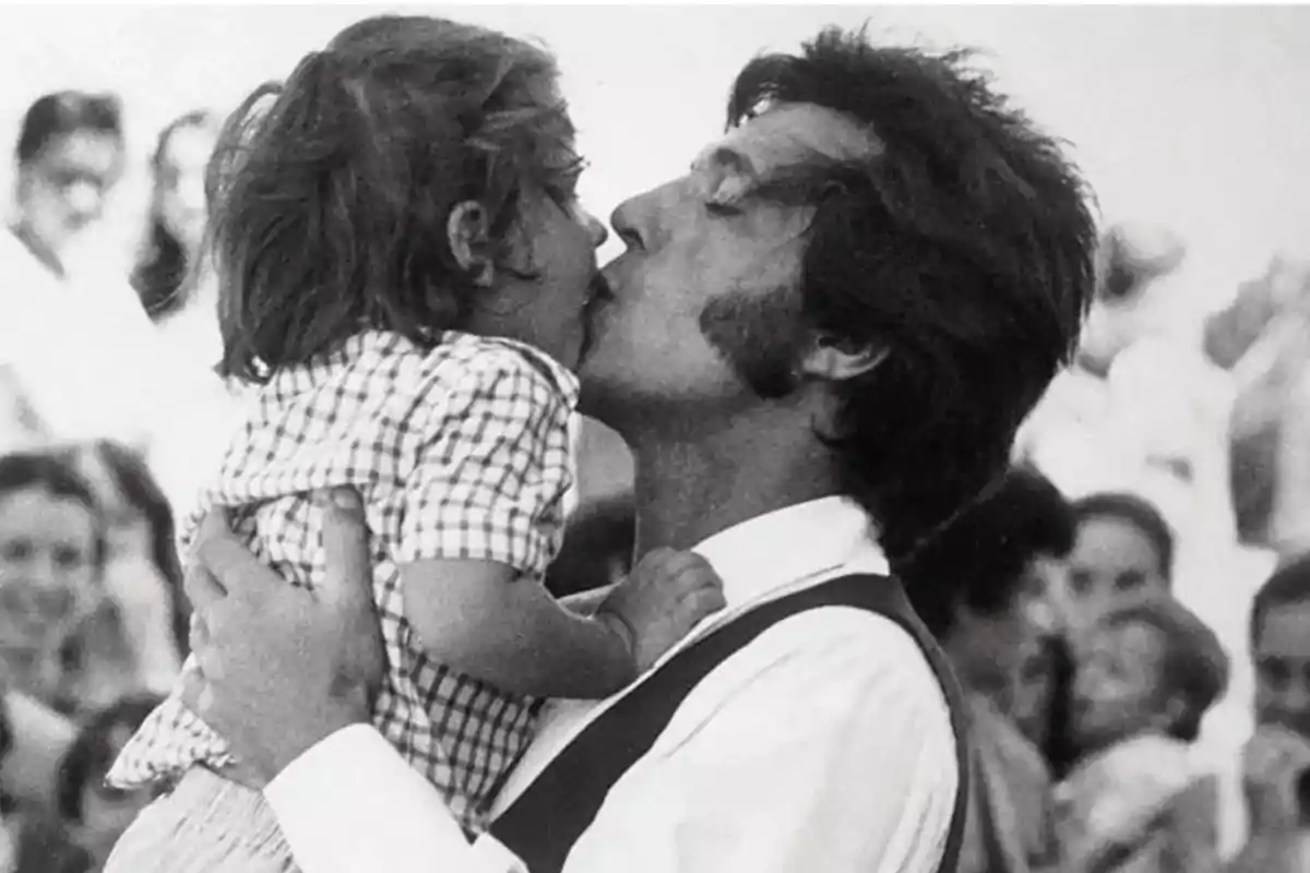 Un hombre besa a una niña pequeña mientras la sostiene en brazos, con personas desenfocadas al fondo.