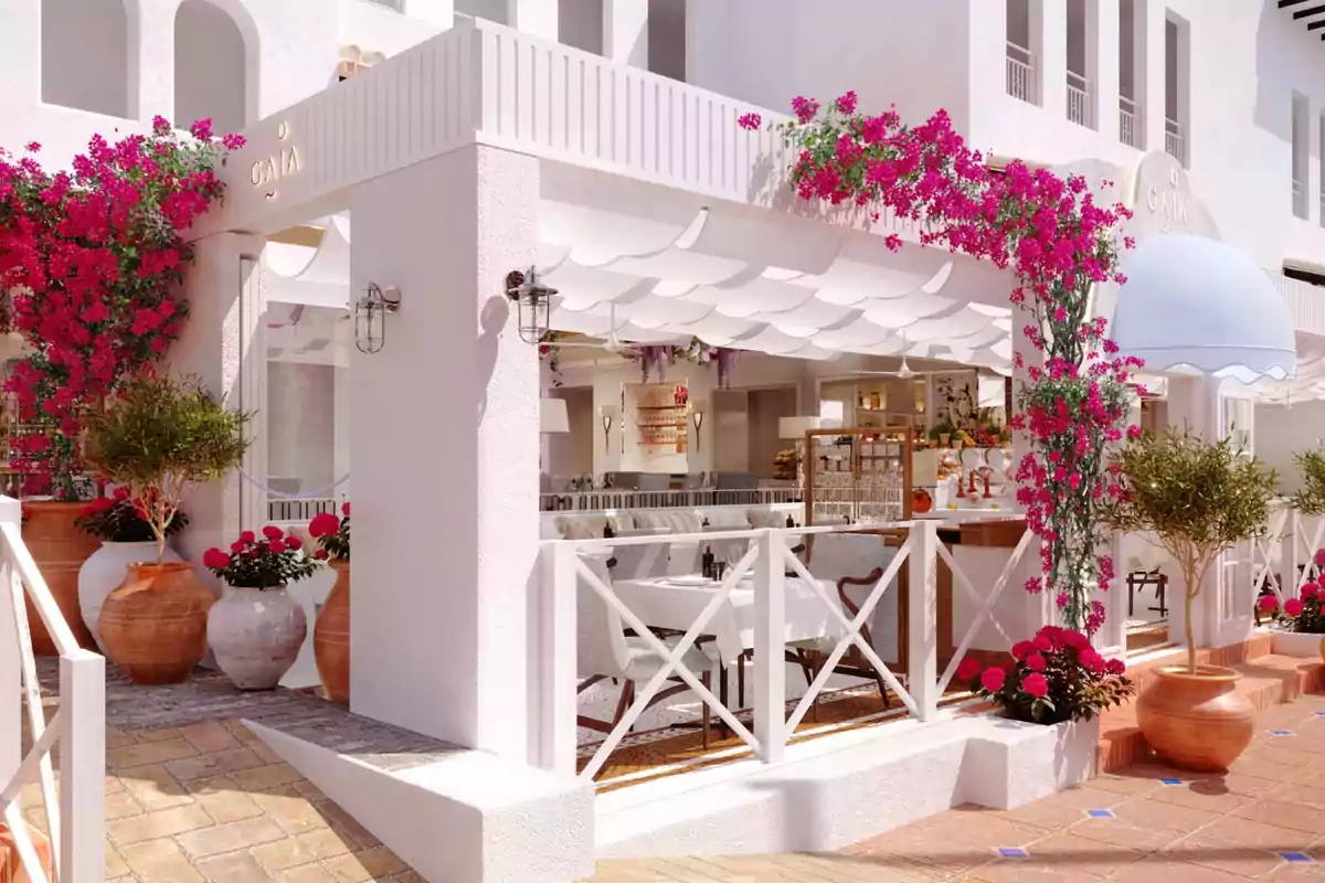 Una terraza de restaurante con decoración mediterránea, flores coloridas y macetas de barro, con mesas y sillas blancas bajo un toldo ondulado.
