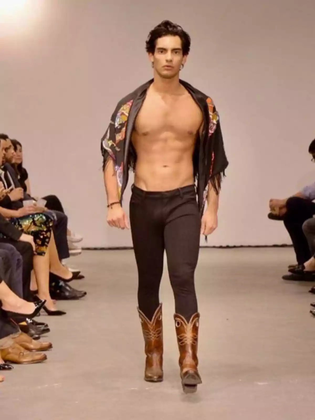 Un modelo masculino camina por la pasarela vistiendo pantalones ajustados negros, botas vaqueras marrones y una chaqueta abierta que deja al descubierto su torso.