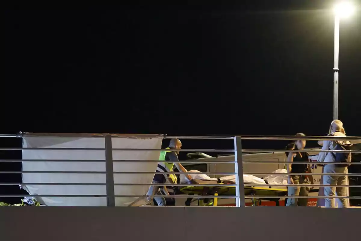 Personas trabajando en una escena nocturna con una camilla y una lona blanca en un puente iluminado por una farola.