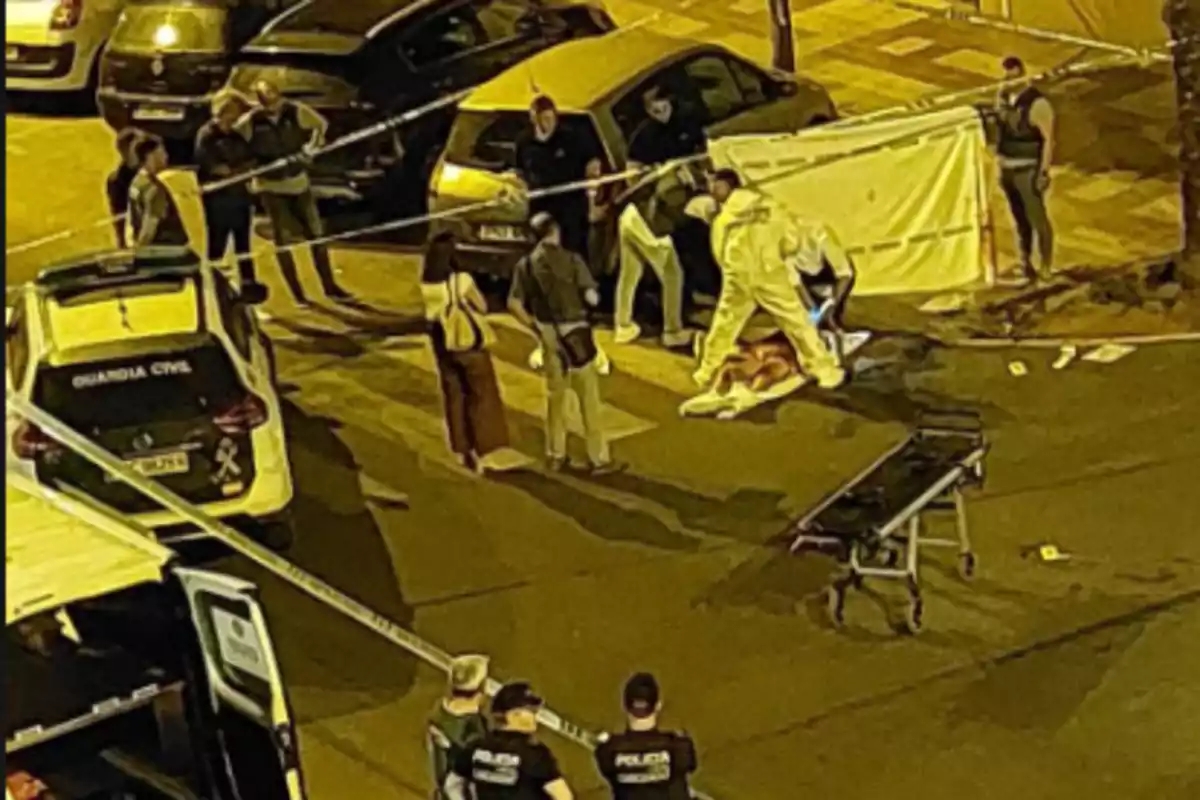 La imagen muestra una escena nocturna de un crimen acordonada por la policía, con varios agentes y personal forense trabajando en el lugar, mientras un cuerpo yace en el suelo cubierto parcialmente por una sábana blanca.