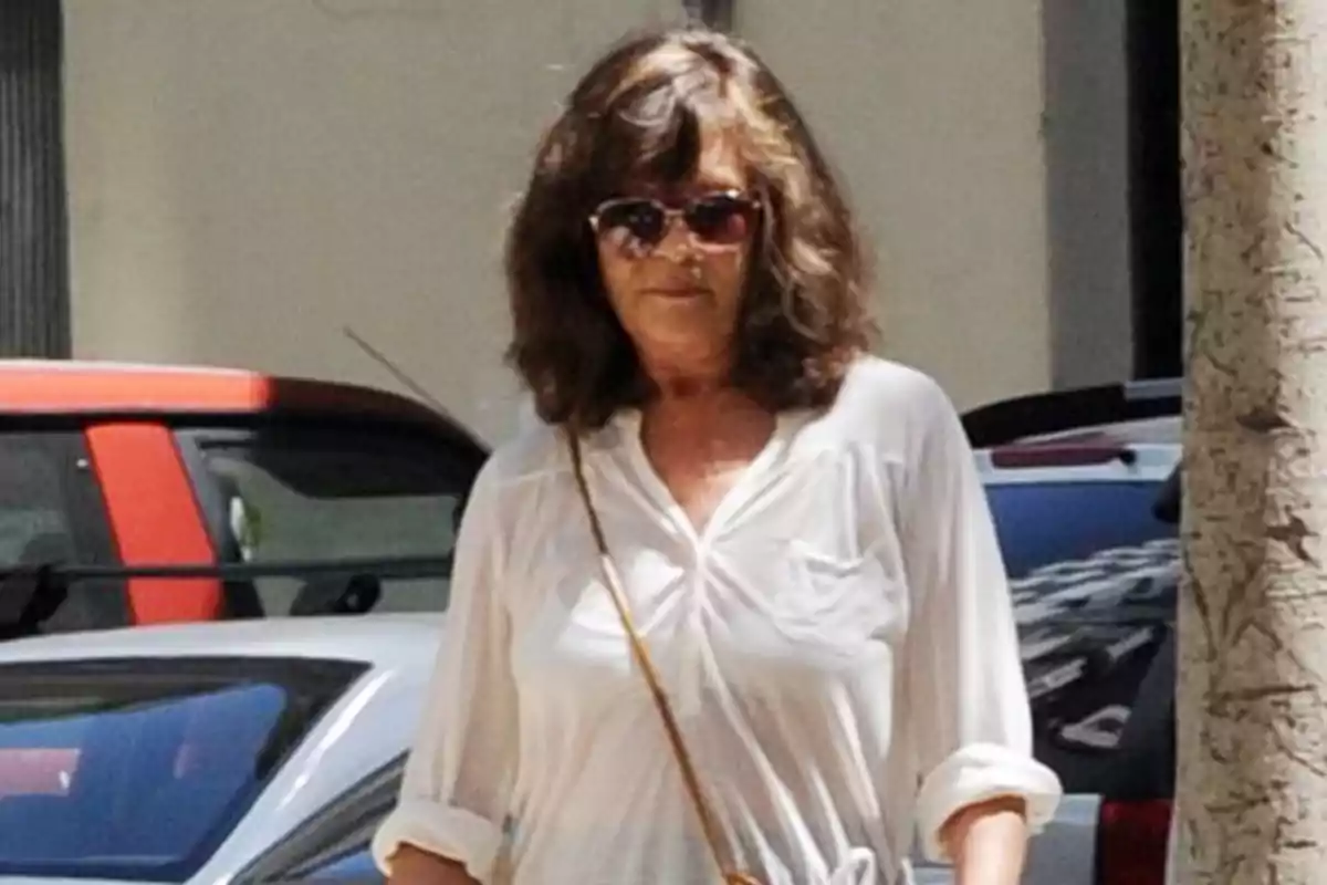 Mujer con gafas de sol y camisa blanca caminando por la calle junto a un coche rojo.