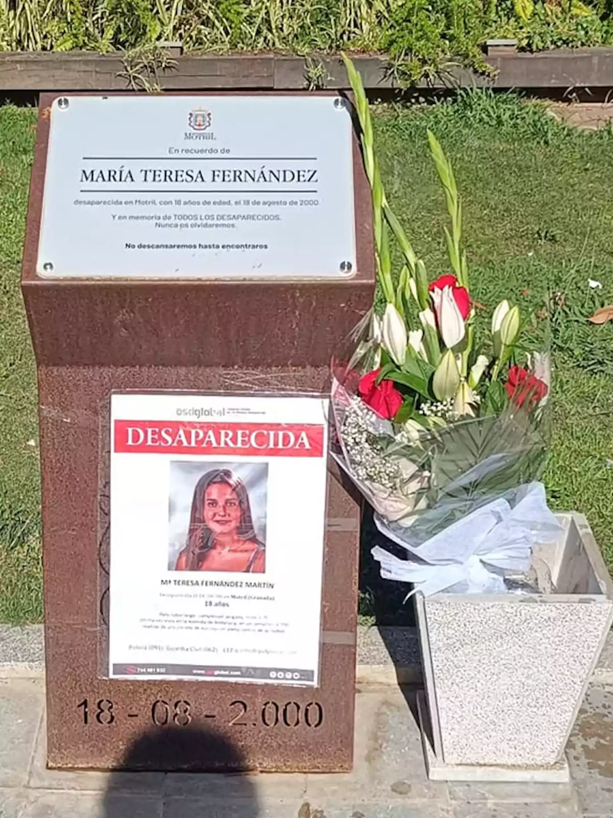 Una placa conmemorativa en honor a María Teresa Fernández, desaparecida el 18 de agosto de 2000, acompañada de un cartel de búsqueda y un ramo de flores.