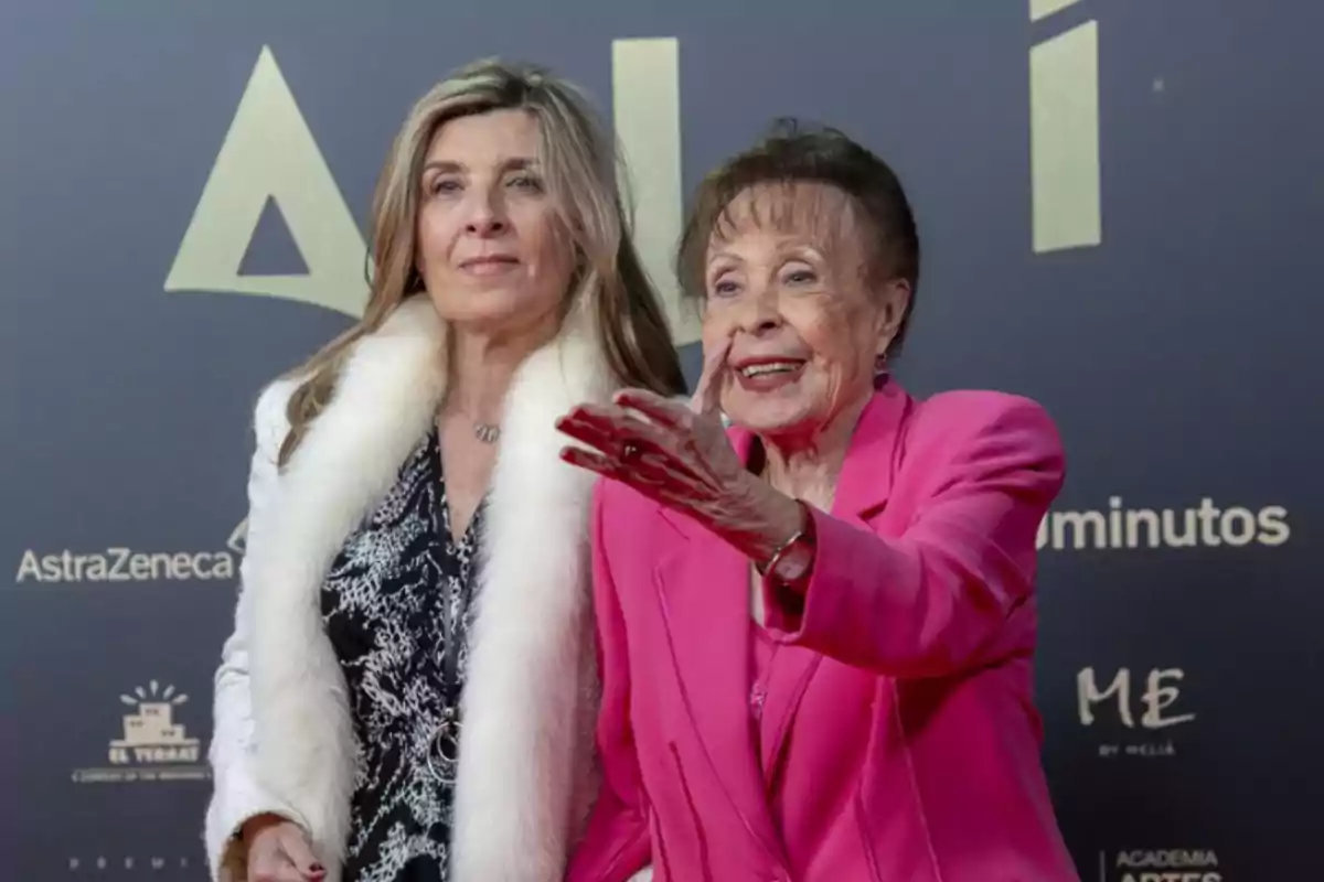 Dos mujeres posan juntas en un evento, una de ellas lleva un abrigo blanco con detalles de piel y la otra un traje rosa.