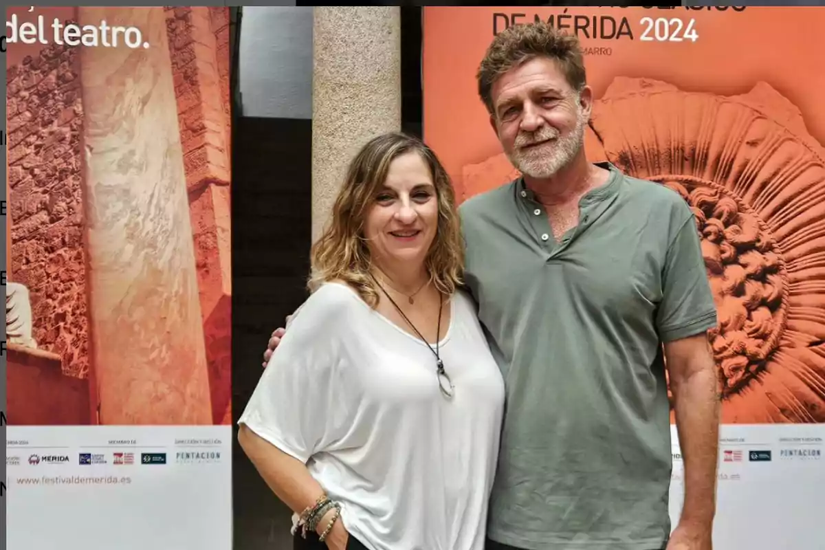 Dos personas posan juntas frente a carteles promocionales del Festival de Mérida 2024.