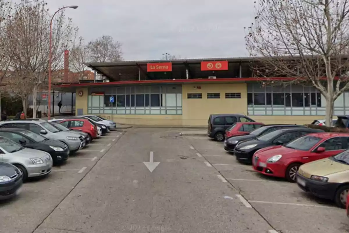 La imagen muestra un estacionamiento lleno de autos frente a una estación de tren llamada 