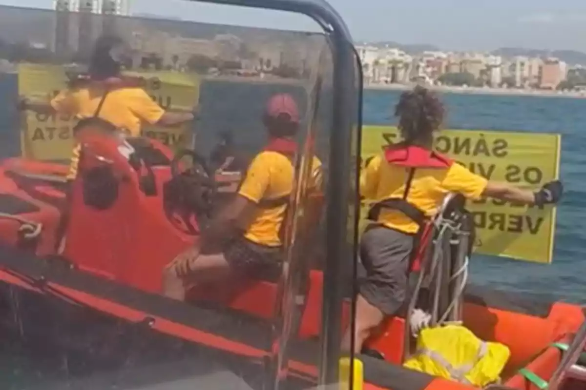 Personas en una lancha inflable sosteniendo carteles amarillos en el mar con una ciudad al fondo.