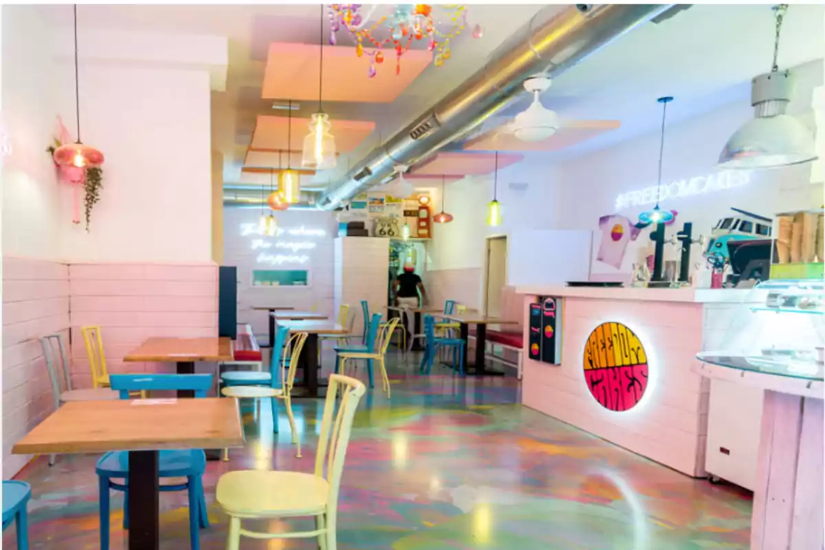 Una cafetería moderna y colorida con decoración vibrante, mesas y sillas de diferentes colores, luces colgantes y un letrero de neón que dice 