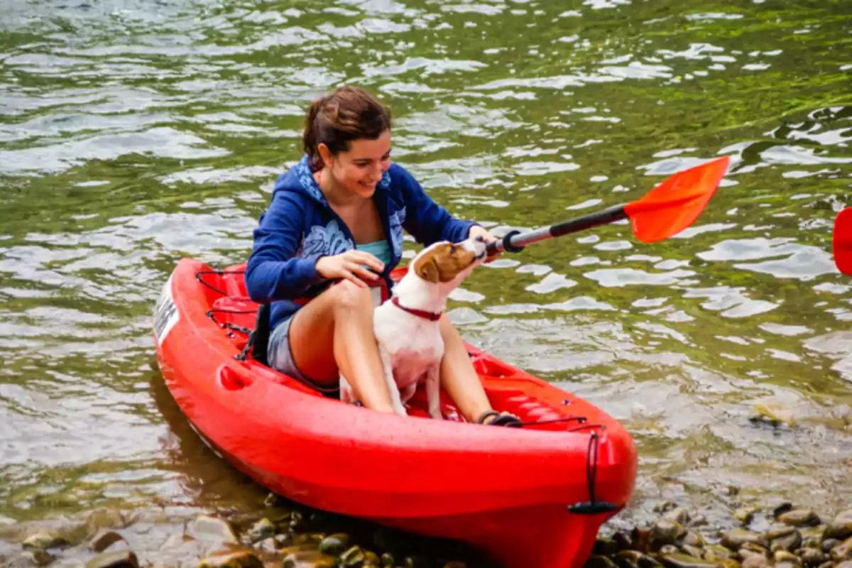 Una mujer en un kayak rojo en el agua, acompañada de un perro que muerde el remo.