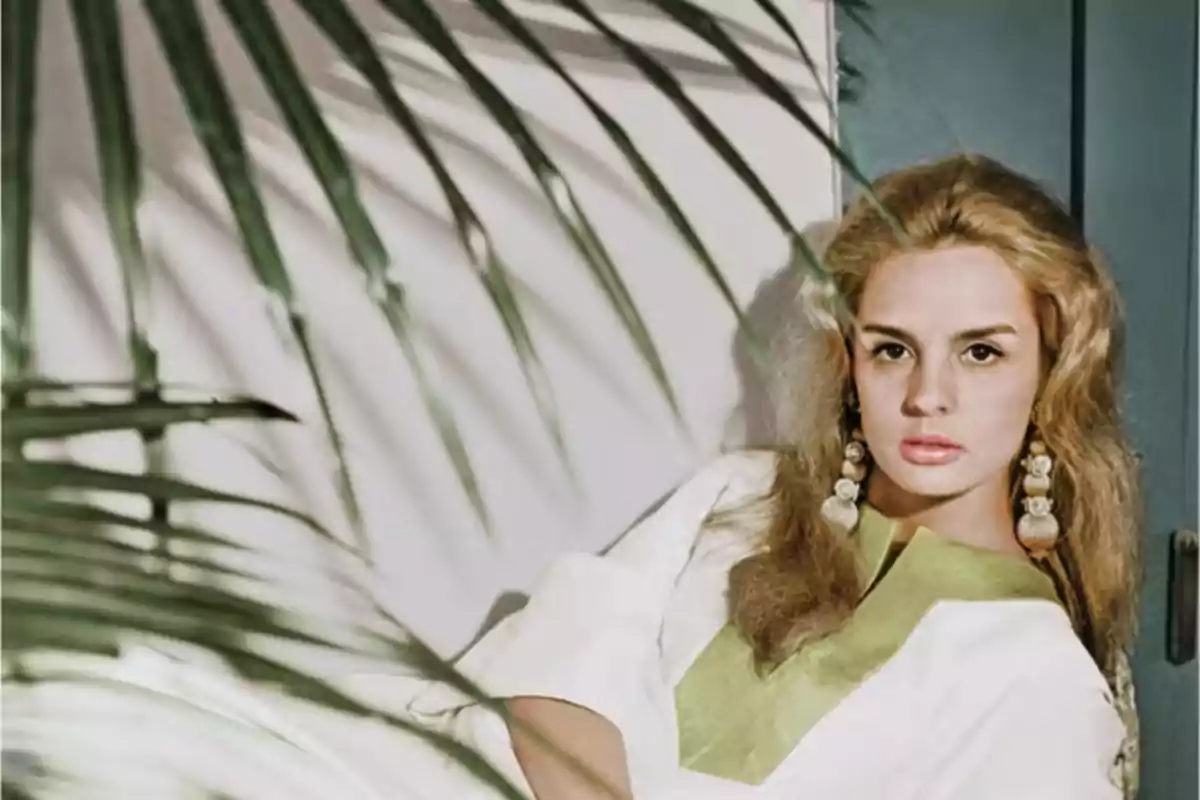 Una mujer con cabello rubio y aretes grandes, vestida con una blusa blanca con detalles verdes, está sentada frente a una pared blanca con sombras de hojas de palmera.