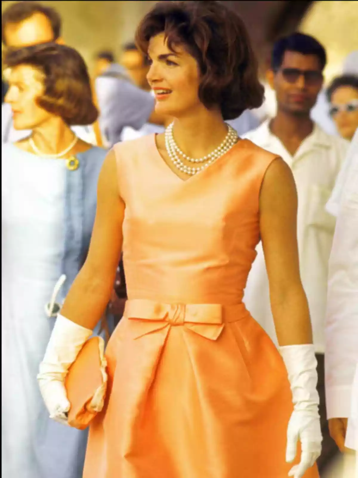 Una mujer con un vestido naranja, guantes blancos y un collar de perlas, sosteniendo un bolso a juego, está de pie junto a otras personas en un evento al aire libre.