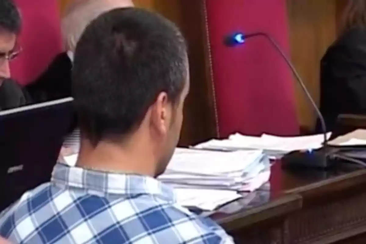 Un hombre de espaldas con camisa de cuadros sentado en una sala de audiencias frente a una mesa con documentos y un micrófono.