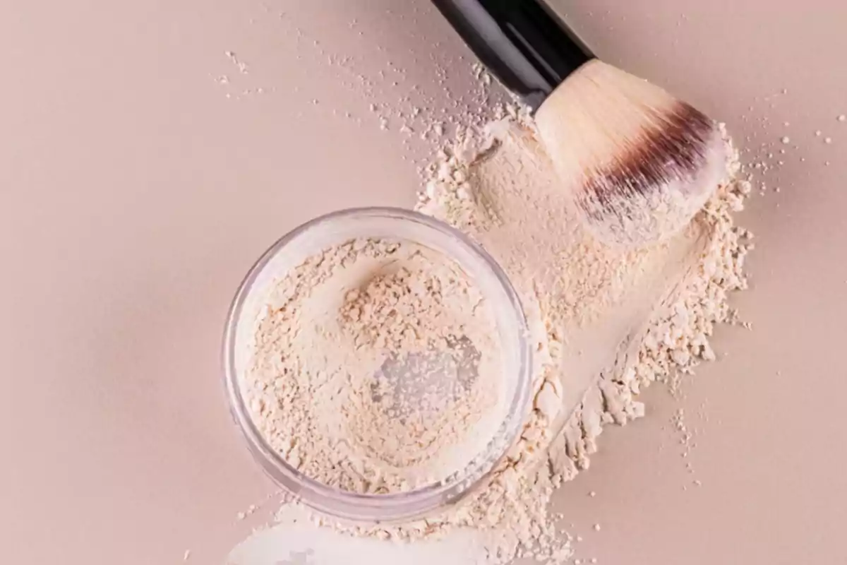 Polvo suelto de maquillaje derramado junto a una brocha sobre una superficie beige.