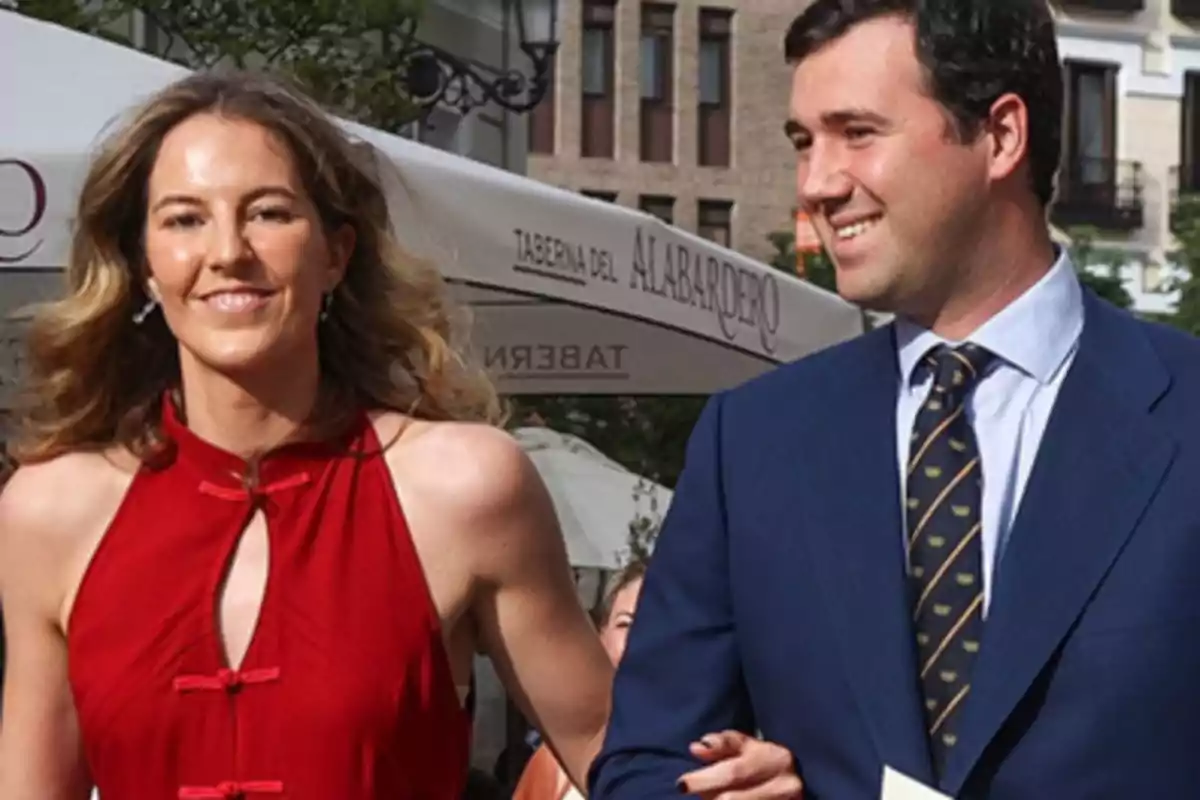 Una pareja sonriente camina al aire libre, ella lleva un vestido rojo sin mangas y él un traje azul con corbata.