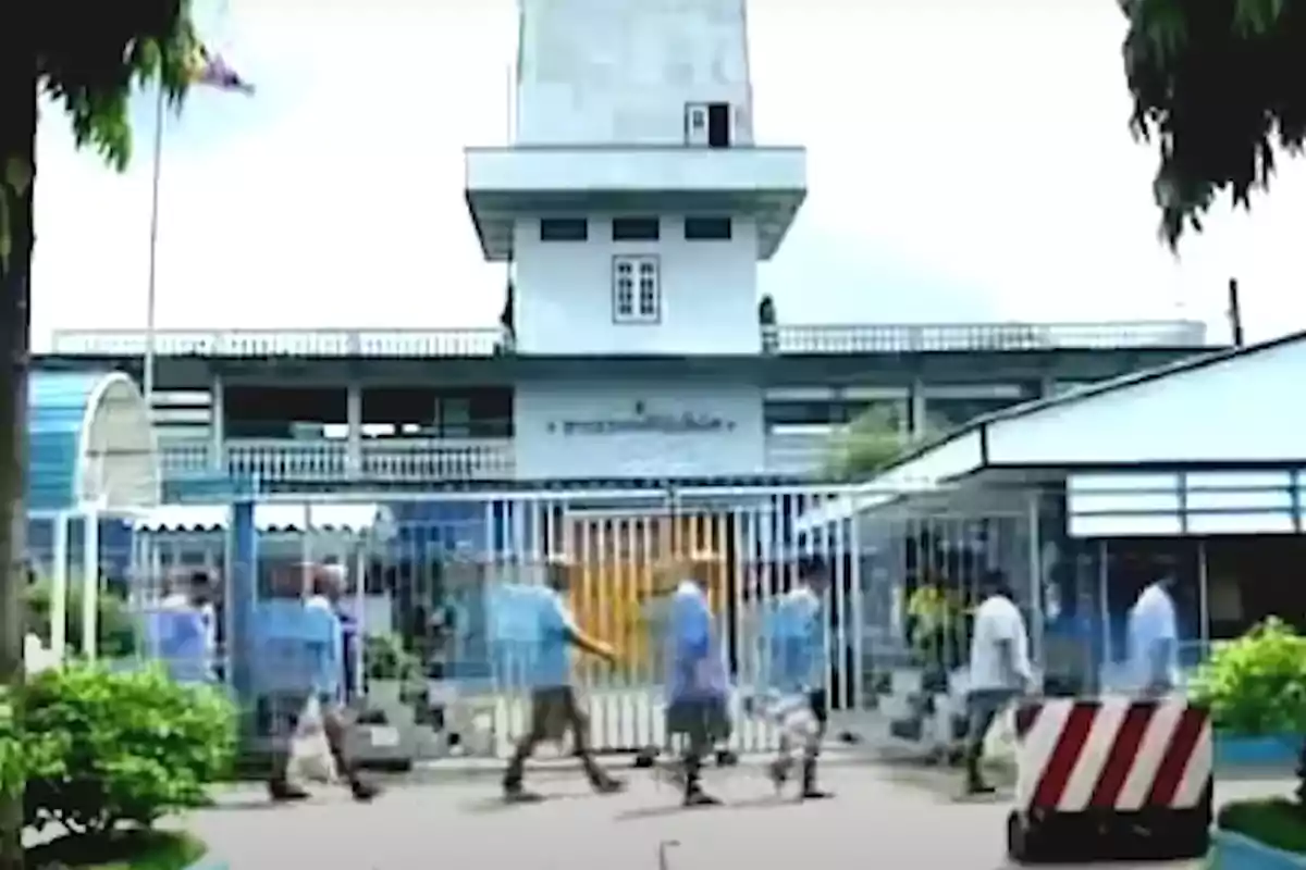 Personas caminando frente a un edificio con una torre y una reja en primer plano.