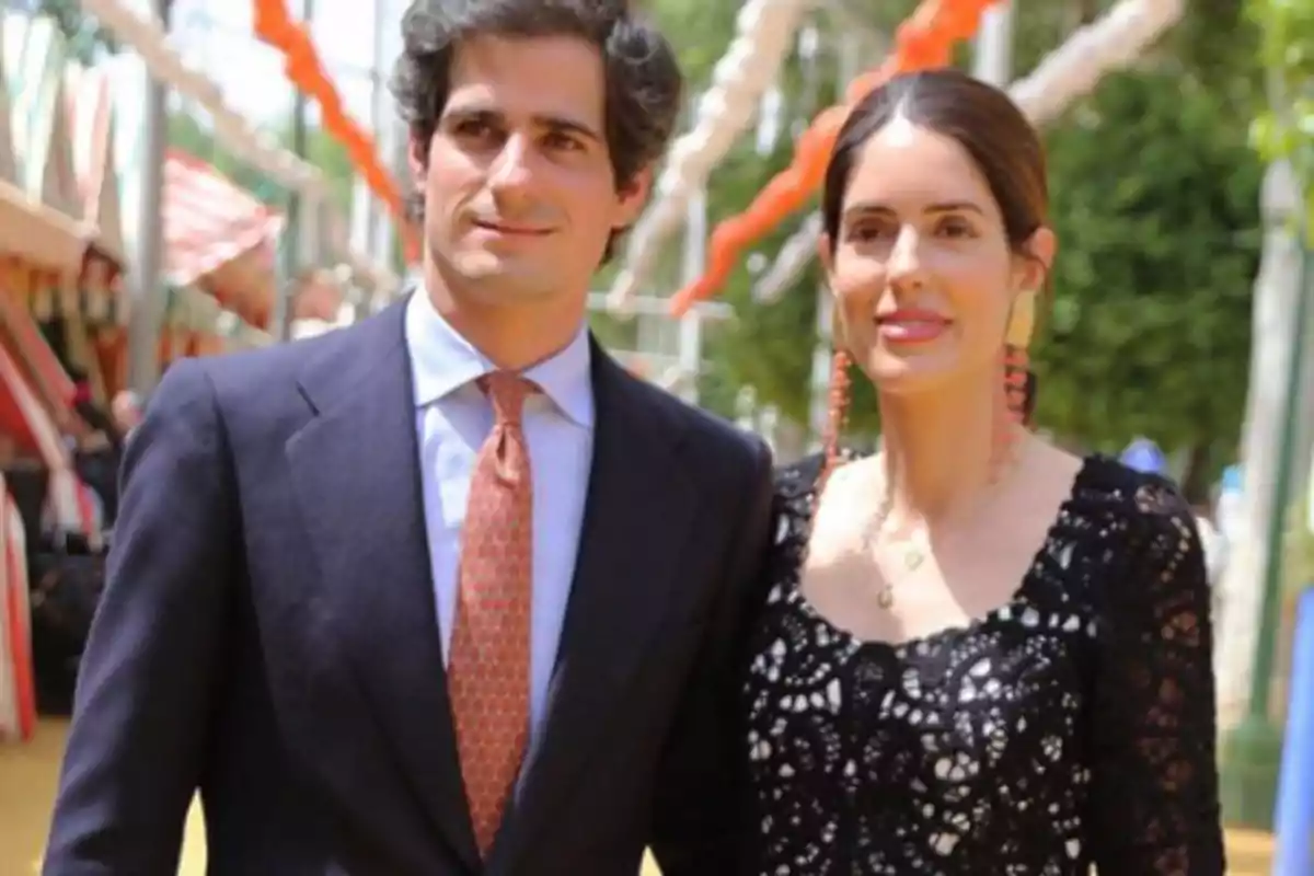 Una pareja elegantemente vestida posa al aire libre en un evento festivo con decoraciones coloridas en el fondo.