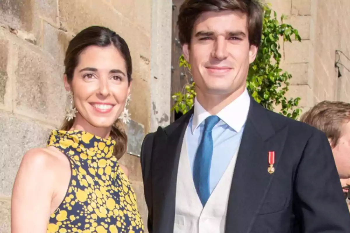 Una pareja sonriente posando juntos en un evento formal, con la mujer usando un vestido estampado y el hombre vistiendo un traje con corbata azul.