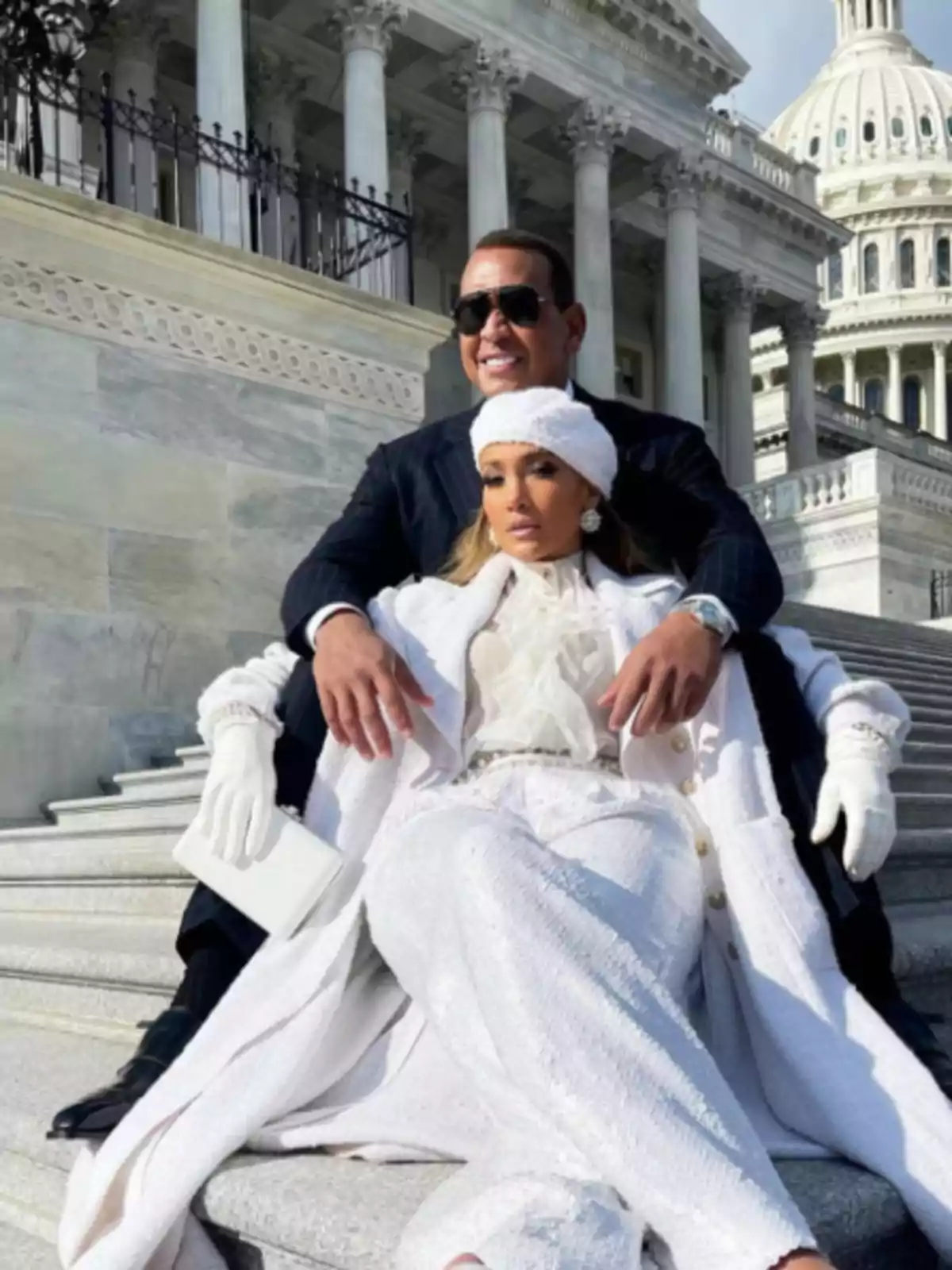 Una pareja elegantemente vestida sentada en las escaleras de un edificio gubernamental con columnas y una cúpula en el fondo.