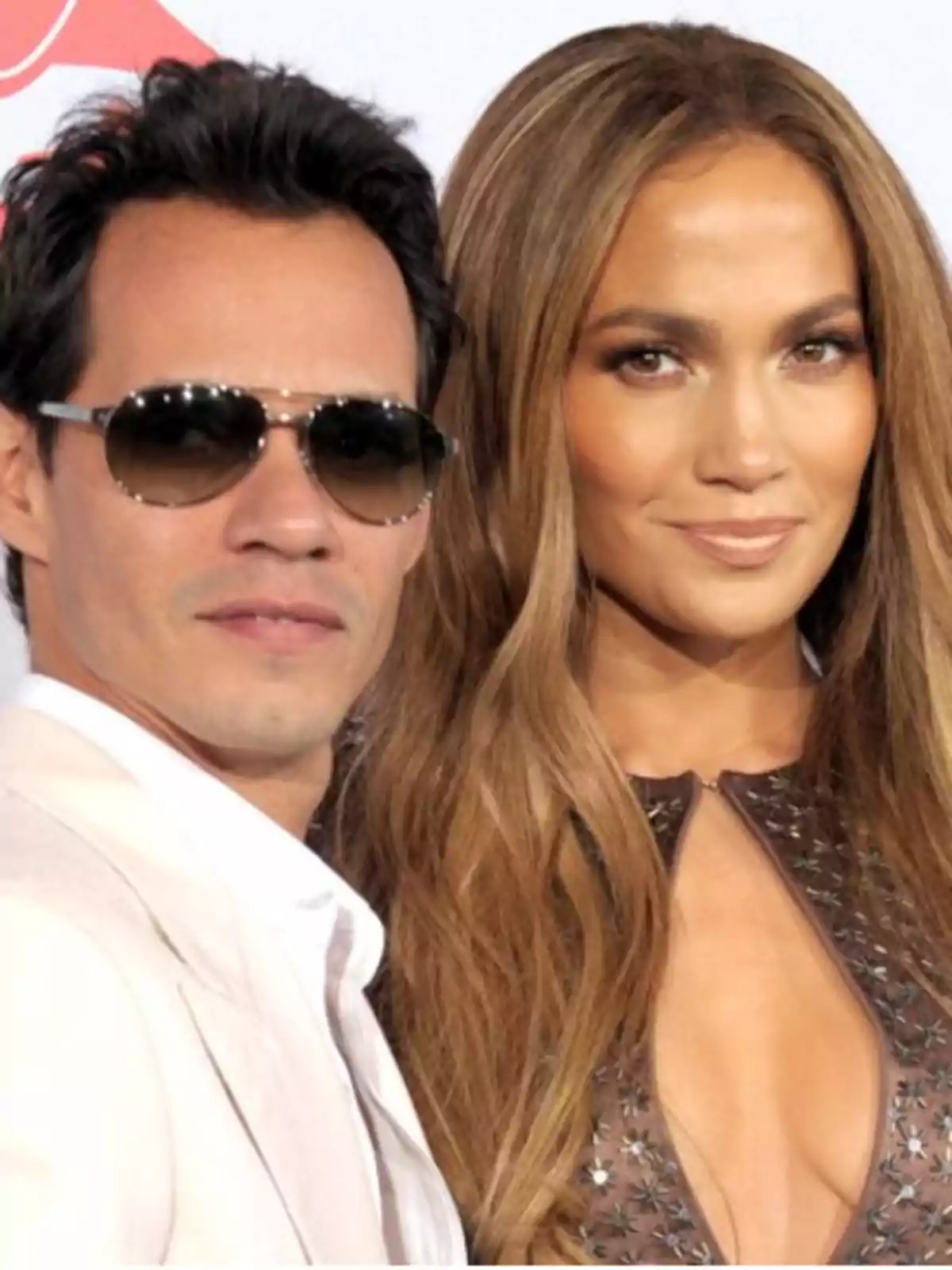 Una pareja posando para una foto, el hombre lleva gafas de sol y un traje claro, mientras que la mujer tiene el cabello largo y suelto y viste un vestido con detalles brillantes.