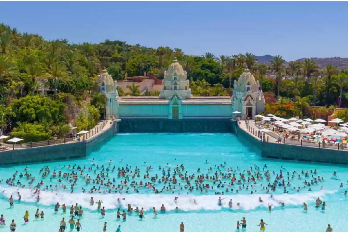 Piscina de olas en un parque acuático con muchas personas disfrutando del agua y rodeada de vegetación y estructuras arquitectónicas.