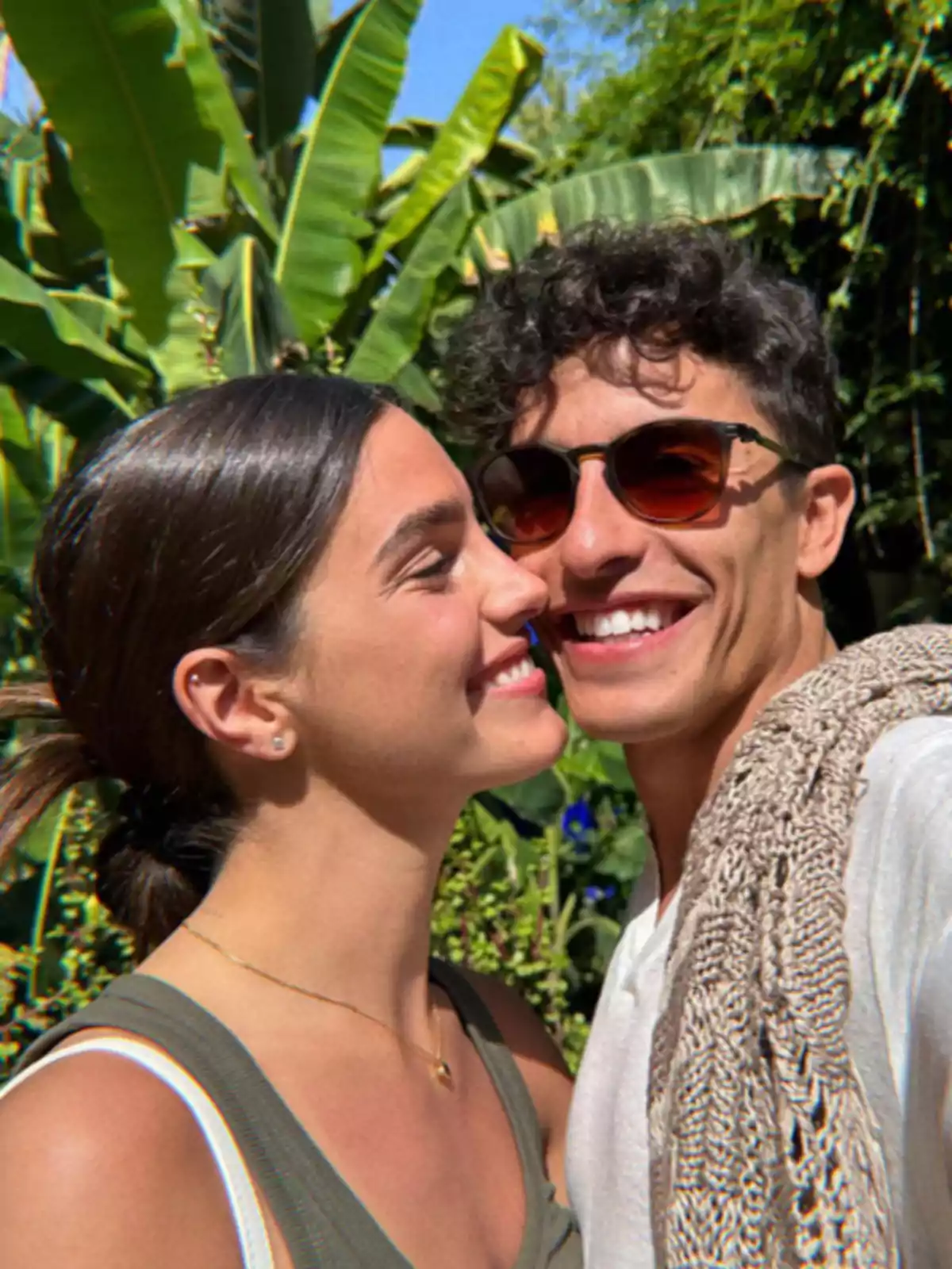 Una pareja sonriente se toma una selfie al aire libre con plantas verdes de fondo.