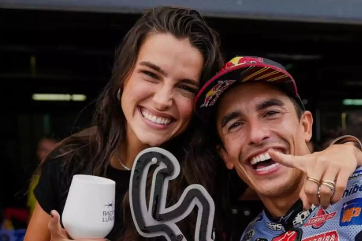 Dos personas sonrientes posando para una foto, una de ellas sostiene una copa y la otra señala hacia la cámara.