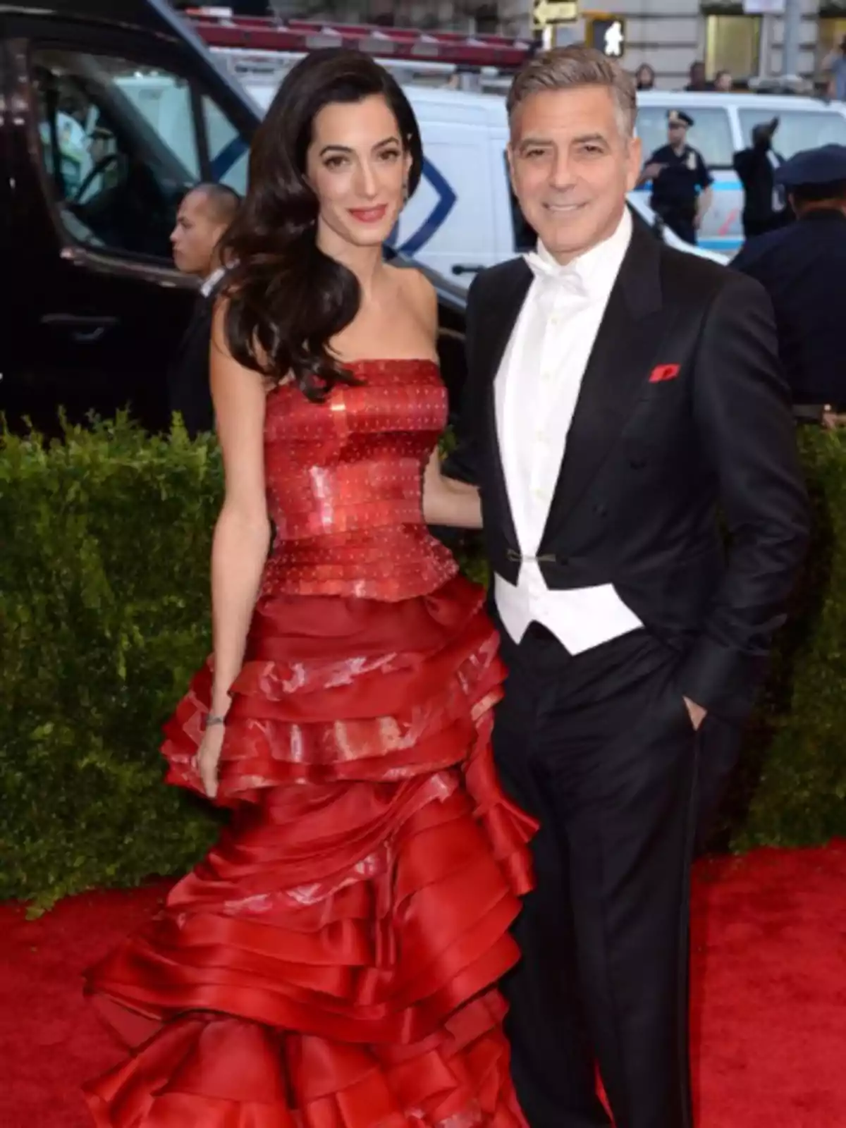 Una pareja elegantemente vestida en un evento de alfombra roja, la mujer lleva un vestido rojo con volantes y el hombre un esmoquin negro con pajarita blanca.