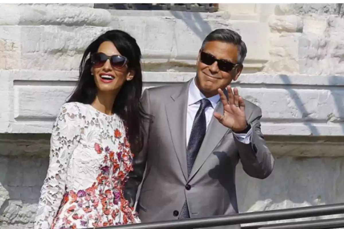 Una pareja sonriente con gafas de sol, el hombre viste un traje gris y la mujer un vestido blanco con detalles florales, ambos están al aire libre frente a una pared de piedra.