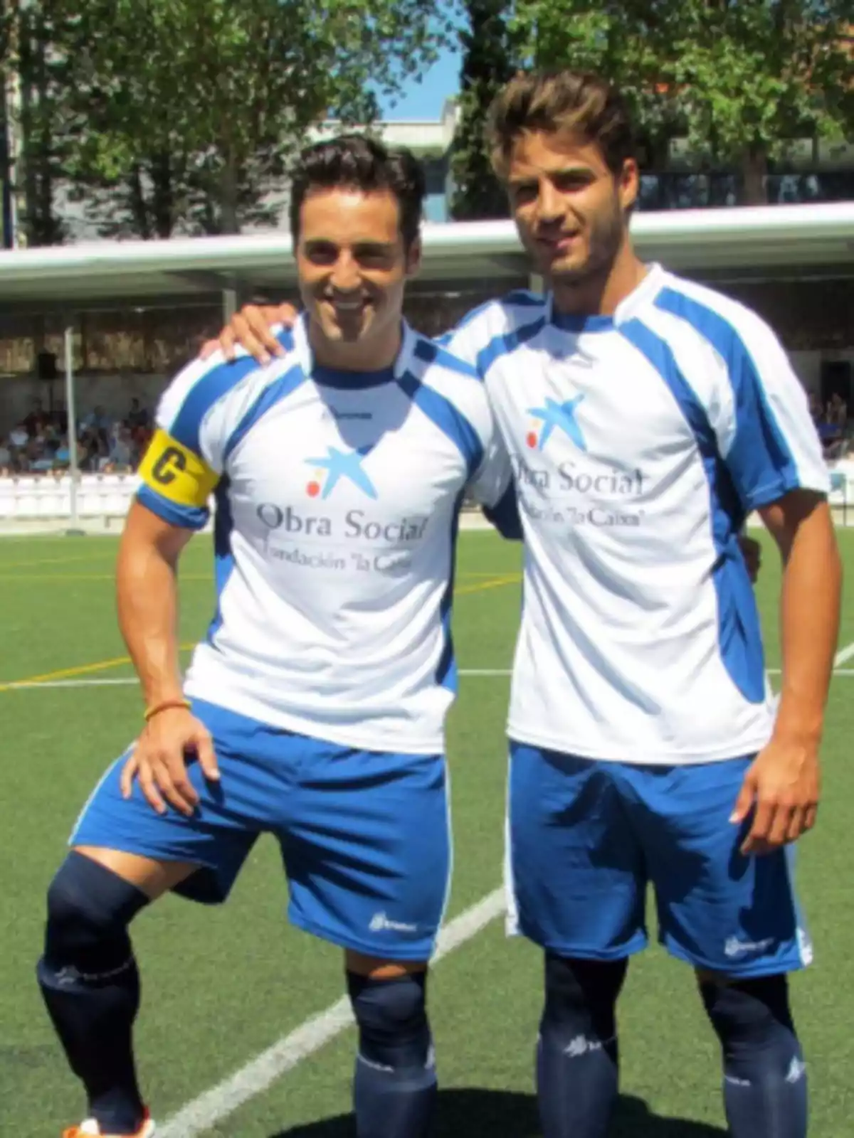 Dos jugadores de fútbol posando en un campo con uniformes blancos y azules de la Fundación 