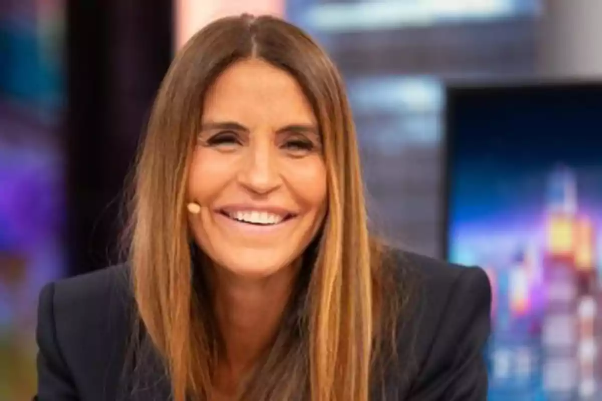 Mujer sonriente con cabello largo y lacio, usando un micrófono de diadema, en un entorno de estudio de televisión.