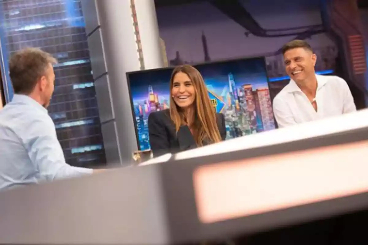 Tres personas conversan y ríen en un set de televisión moderno con un fondo de ciudad iluminada.