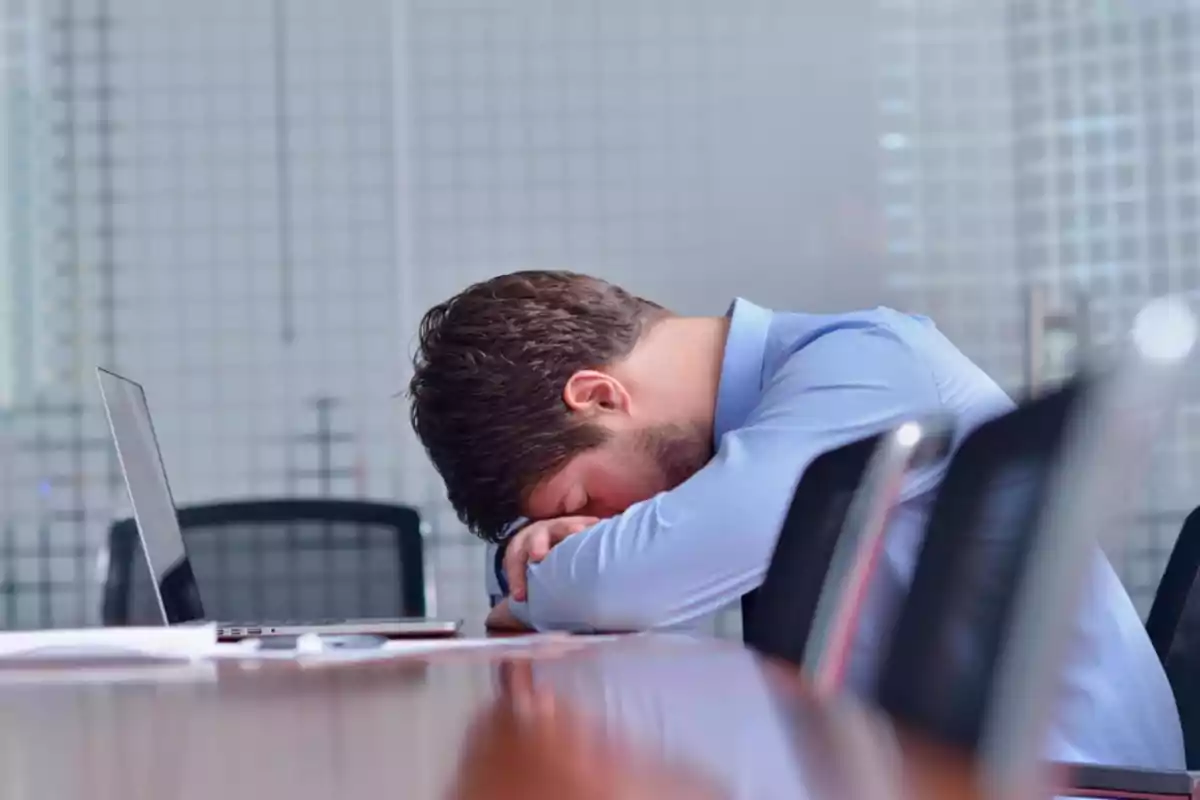 Hombre durmiendo sobre una mesa en una sala de reuniones con una computadora portátil abierta.