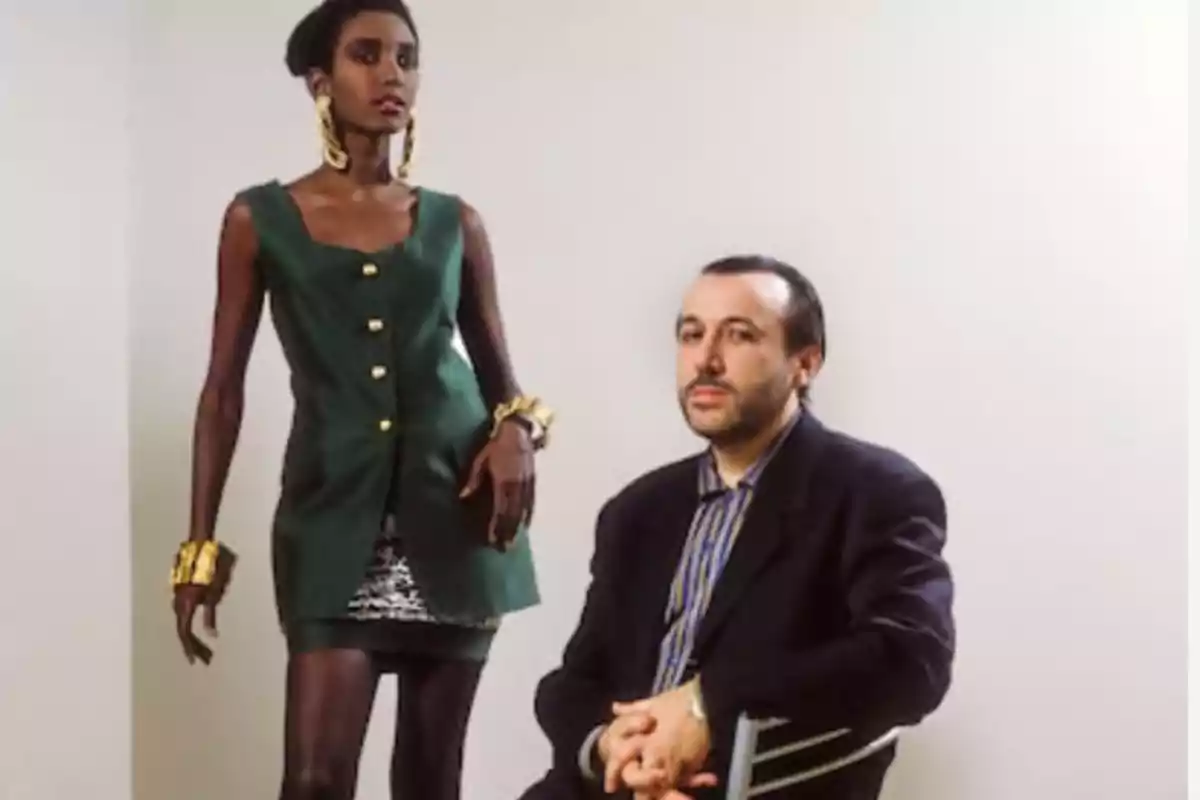 Una mujer de pie con un vestido verde y joyas doradas junto a un hombre sentado en una silla con traje oscuro.