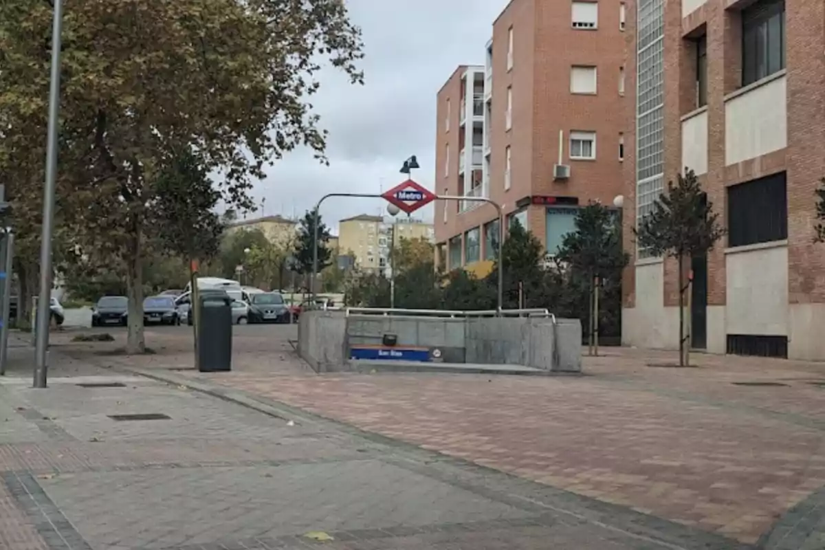 Entrada a la estación de metro San Blas en una zona urbana con edificios de ladrillo y árboles.