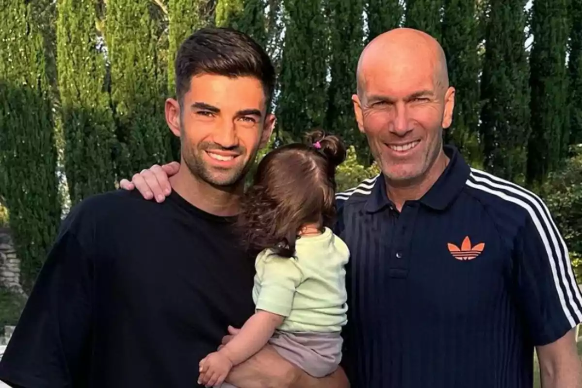 Dos hombres sonrientes posan al aire libre con un bebé en brazos, con árboles altos de fondo.