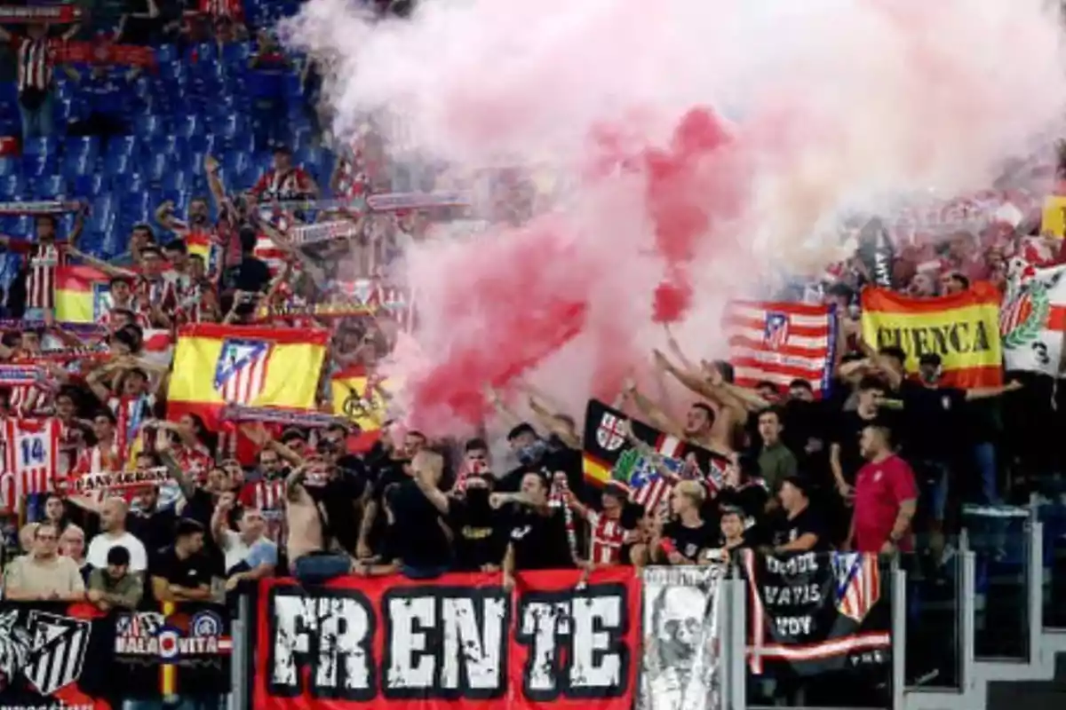 Aficionados de un equipo de fútbol celebrando en las gradas con banderas, bufandas y bengalas de humo rojo.