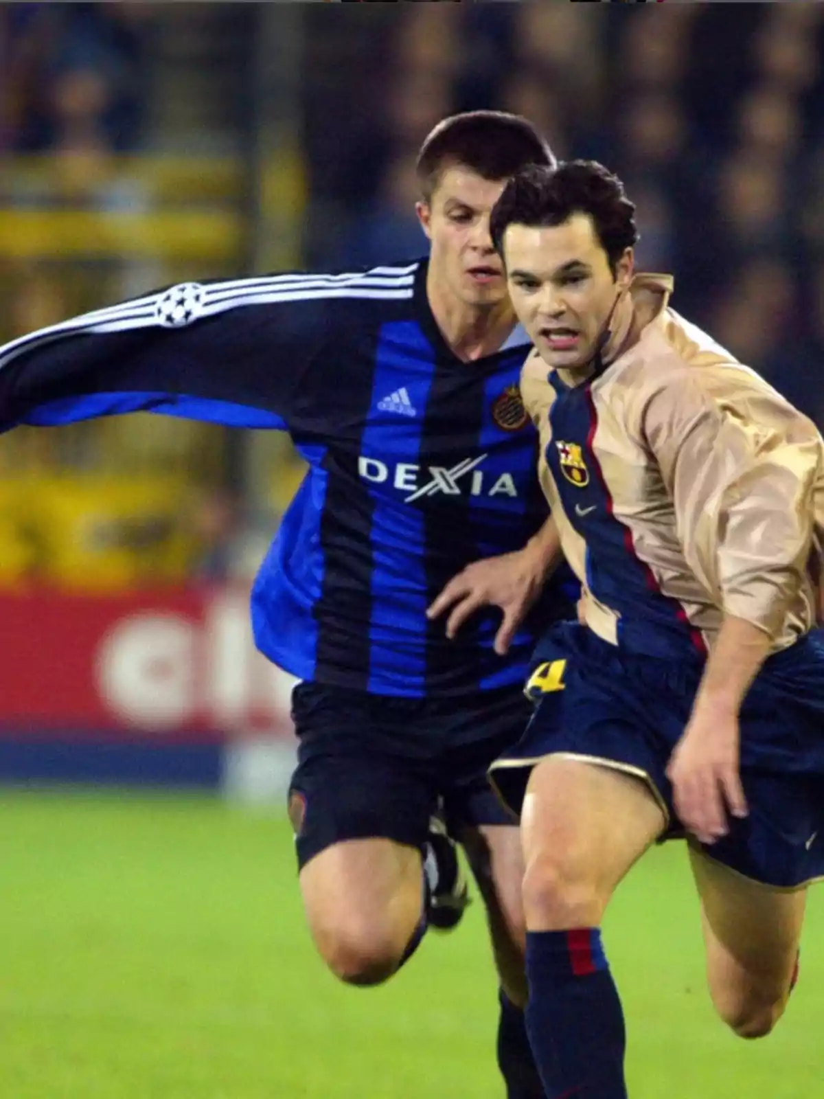 Dos jugadores de fútbol compiten por el balón en un partido, uno con uniforme azul y negro y el otro con uniforme beige y azul.