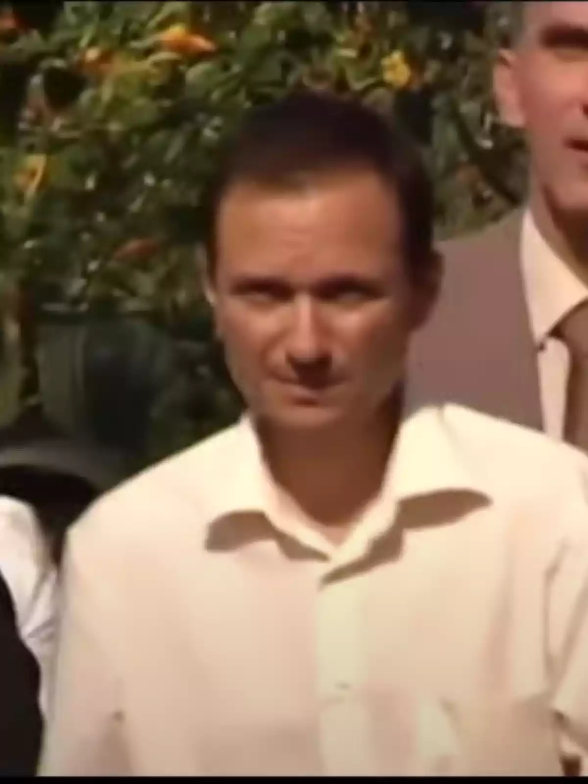 Un hombre con camisa blanca está de pie al aire libre, con vegetación de fondo y otras personas alrededor.