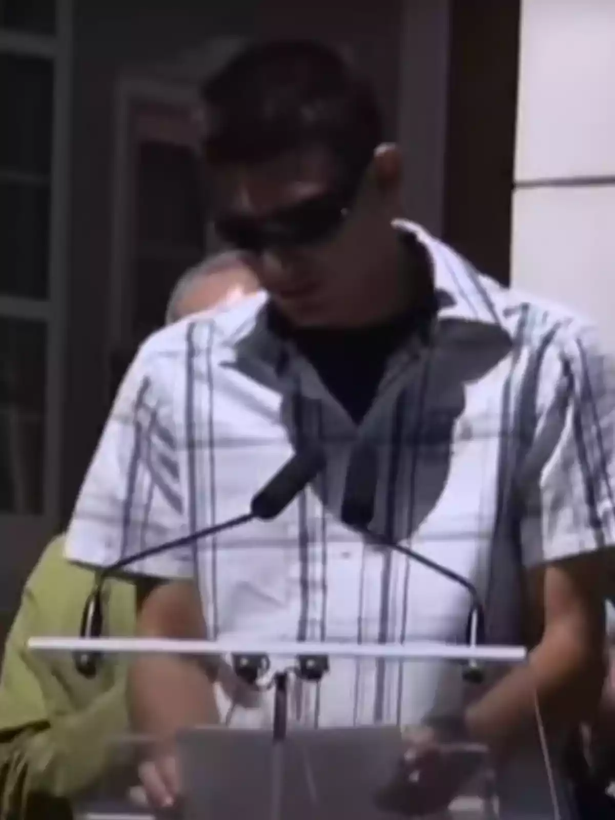 Un hombre con gafas de sol y camisa a cuadros habla frente a un micrófono en un podio.